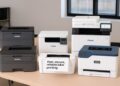 Five laser printers arranged on an office testing table, including Brother, Canon, HP, and Xerox models, used for hands‑on comparison