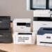 Five laser printers arranged on an office testing table, including Brother, Canon, HP, and Xerox models, used for hands‑on comparison