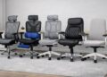 Five ergonomic office chairs — SIHOO M57, FlexiSpot C7DG, Hbada E3 Air, Nouhaus +Posture, and Boulies NUBI — lined up side by side on a light gray carpet in a modern home office with a light wood desk, potted plant, and sheer white curtain, showcasing their different designs and materials.