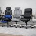 Five ergonomic office chairs — SIHOO M57, FlexiSpot C7DG, Hbada E3 Air, Nouhaus +Posture, and Boulies NUBI — lined up side by side on a light gray carpet in a modern home office with a light wood desk, potted plant, and sheer white curtain, showcasing their different designs and materials.