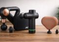 hree massage guns — Theragun Sense, Hypervolt Go 3, and Theragun Mini — arranged on a wooden surface with soft natural lighting