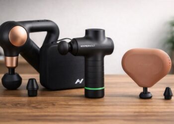 hree massage guns — Theragun Sense, Hypervolt Go 3, and Theragun Mini — arranged on a wooden surface with soft natural lighting