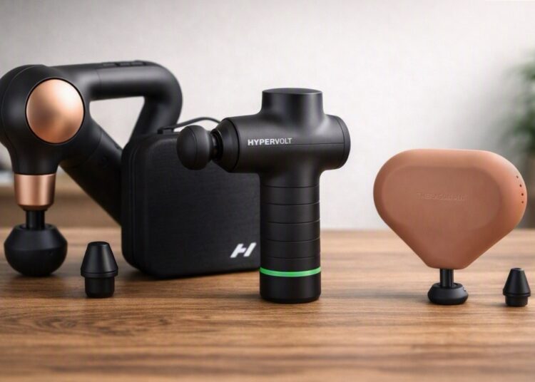 hree massage guns — Theragun Sense, Hypervolt Go 3, and Theragun Mini — arranged on a wooden surface with soft natural lighting