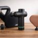 hree massage guns — Theragun Sense, Hypervolt Go 3, and Theragun Mini — arranged on a wooden surface with soft natural lighting