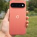 Coral Google Pixel 11 smartphone held outdoors showing rear camera bar and sleek design.