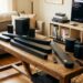 A collection of soundbars, subwoofers, and remotes arranged on a wooden table in a living room setup for hands-on audio testing, with a TV displaying audio test software in the background.