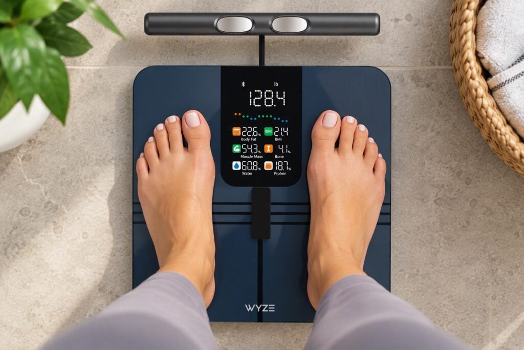 Person standing on a Wyze Scale BodyScan smart scale displaying body composition metrics, weight, BMI, muscle mass, body fat percentage, and wellness tracking data.