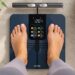 Person standing on a Wyze Scale BodyScan smart scale displaying body composition metrics, weight, BMI, muscle mass, body fat percentage, and wellness tracking data.