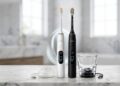White and black Philips Sonicare DiamondClean 9000 electric toothbrushes standing on a marble bathroom countertop next to a white charging puck base and a glass charging base with USB cables